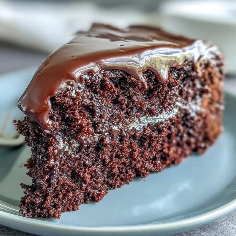 Slice of Easy Chocolate Fudge Cake served on a white plate with fresh raspberries, showcasing the rich, dark crumb and glossy chocolate frosting.