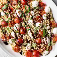 A close-up of Caprese Couscous Salad in a white bowl, featuring glossy balsamic glaze drizzled over mozzarella, tomatoes, and couscous.
